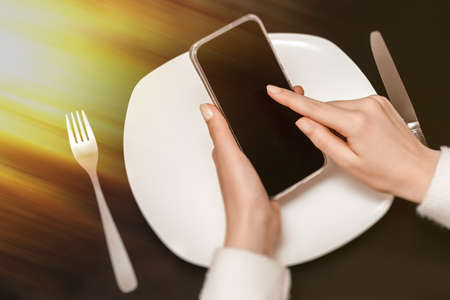 Food delivery concept. Top view on smartphone, hands holding phone showing screen. Smart phone black screen and white plate, fork and knife, black background table. Take away restaurant meal onlineの写真素材