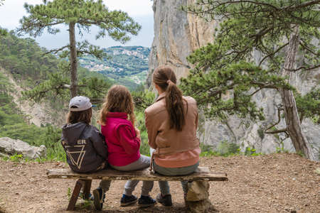 camping, travel, tourism, hike and people concept - children sit on the bench with their backs to us and look at the beautiful landscape in woodsの写真素材
