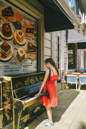 A beautiful girl walks outdoors, dressed in a red sundress, plays the street piano near a cafe, summer timeの写真素材