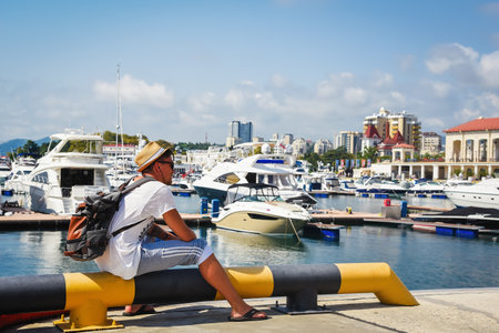 Sochi, Russia, July 27, 2019: traveler man with a backpack on his back sits on the dock and looks at the port. Building of main port of Sochi. summer timeのeditorial素材