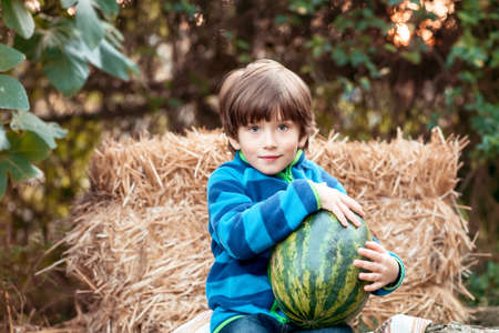 On a sunny autumn day, a cute toddler boy sits near a large watermelons and smiles sweetly. Waiting for a delicious dessertの写真素材