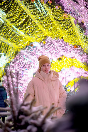 Young smiling happy girl in Christmas decorated with holiday lights. New Year's Eve, festive atmosphere. Moscow, Tverskoy Boulevard. Spending winter time outdoors. Warm clothes, knitted hat, mittensの写真素材
