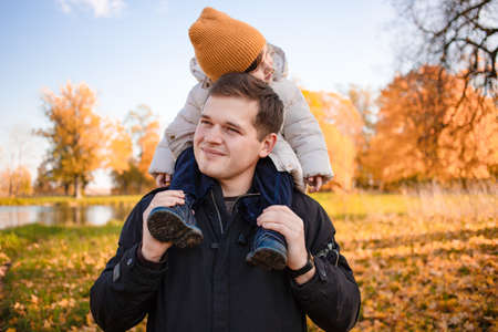 Father gives his son a ride on his back in the park. Portrait of a pensive father giving his daughter a piggyback ride on his shoulders and looking up. Cute baby girl with her fatherの写真素材