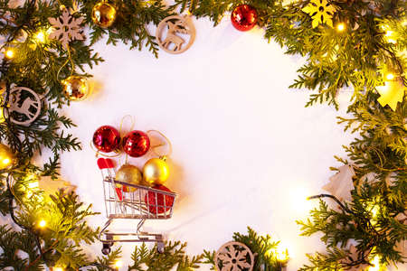 Christmas or New Year background: fir branches on a white background with wooden toys and a sparkling garland, bells, balls. White frame for congratulations. Shopping basket with christmas ballsの写真素材