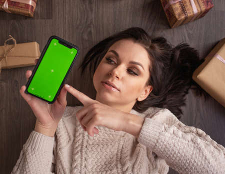 Young emotional brunette woman lies and shows smartphone with green copy space screen, focus on screenの写真素材
