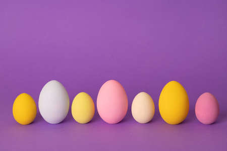 A row of colorful Easter eggs painted in simple colors on a light purple background - side view with...
