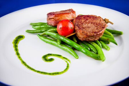 Fried pork medallions on skewers with string beans and one whole tomato. Closeup on a white plate on a blue background. Ketogenic diet - high-fat nutrition. Paleo Diet. Pegan diet.の写真素材