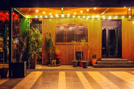 Summer terrace in the cafe in the evening, decorated with garlands, with plants in pots, barrels on tiled floor. Menu board on the wall. Light bulb garlandの写真素材