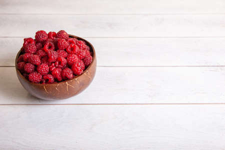 Raspberries in a coconut shell bowl on a white wooden background.