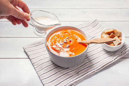 Woman pours out the cream in Pumpkin soup-puree with breadcrumbs and seeds, white wooden background.の写真素材