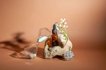 Modern still life with serum bottle on background of unusual natural stone, with bouquet of lilies of the valley and hourglass. The concept of anti-aging care products.の写真素材