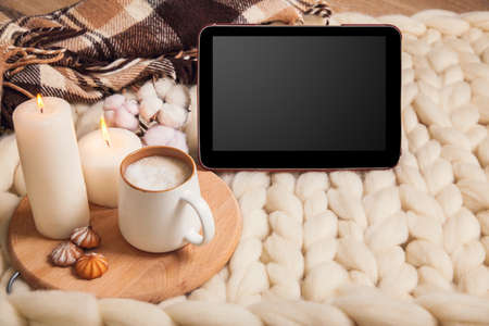 Mockup tablet with Cup of cappuccino and cookies, candles, checkered plaid on the background of blanket of thick yarn. The atmosphere of homeliness and comfort.の写真素材