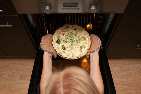 Woman takes an open shortbread pie with chicken and broccoli, sprinkled with cheese out of the oven in her kitchen.の写真素材