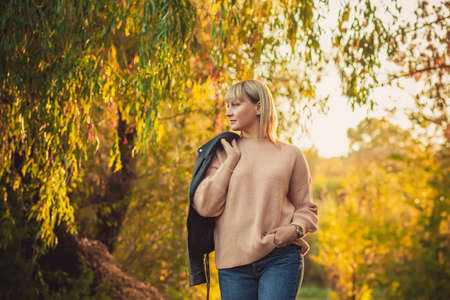 A blonde woman with a short haircut walks through the forest in a knitted sweater and a leather jacket on her shoulder. Outdoor hiking in autumn forestの写真素材