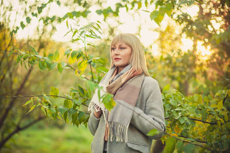 A blonde woman with a short haircut walks through the forest in a gray wool coat and a plaid scarf. Walking in nature in autumn forestの写真素材
