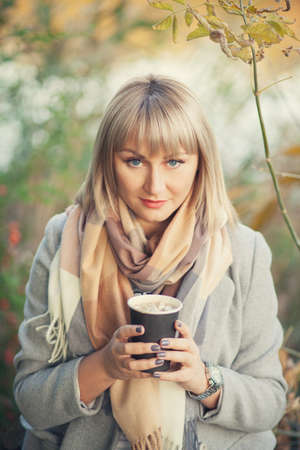 Blonde woman with short haircut in gray wool coat and plaid scarf drinks coffee cappuccino with marshmallows. Walking and picnic in nature in autumn forestの写真素材