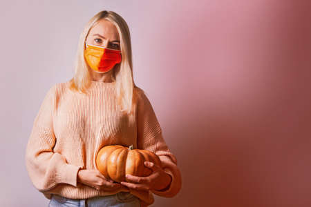 Unhappy young blonde woman wearing medical mask in warm sweater hold pumpkin.の写真素材