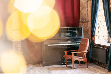 Old vintage piano and armchair stands in the hall against the background of a red curtain and a brick wall near the big window, with bokeh and highlights.の写真素材