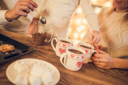 Family: mom, dad and daughter in white sweaters cook and drink cocoa with marshmallows. Closeup hands and cups. Christmas concept.の写真素材