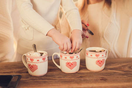 Family: mom, dad and daughter in white sweaters cook and drink cocoa with marshmallows. Closeup hands and cups. Christmas concept.の写真素材