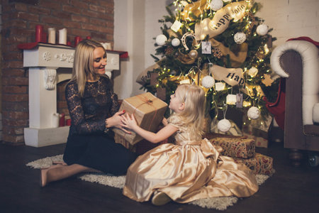 A little girl in an elegant dress gives a mom a gift under the Christmas tree. Mother and daughter with gift box.の写真素材