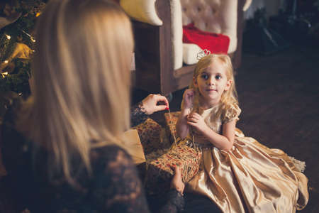 A little girl in an elegant dress and her mom open a gift box under the Christmas tree.の写真素材