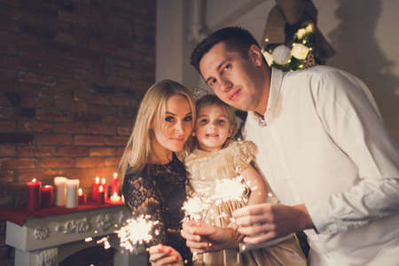 Happy family - mother, father and daughter holding sparklers on the background of a Christmas tree and fireplace with candles.の写真素材