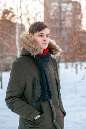 A young man in a winter jacket with fur stands in a park against the background of a snow-covered city.の写真素材