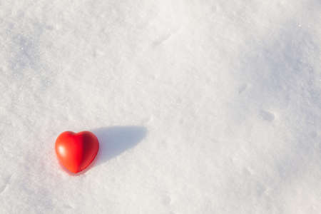 The red heart of the rubber antistress toy lies on the snow cover in sunny winter evening on sunset. Minimalistic snow background with heart, copy spaceの写真素材