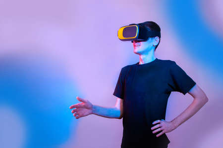 A young man in virtual reality glasses gives a hand to say hello. Communication in virtual reality. Neon colors, copy spaceの写真素材