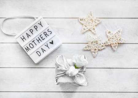 Gift box trendy wrapped in silk fabric in Furoshiki technique, white flower Eustoma or Lisianthus, light box with lettering Happy Mother's Day on light wooden background.の写真素材