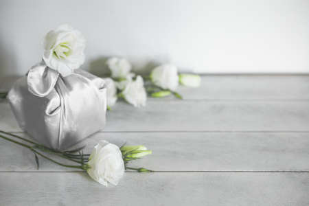 Gift box wrapped in silk fabric in Furoshiki technique, white flowers Eustoma, light wooden background with copy space. Zero Waste Life Concept.の写真素材