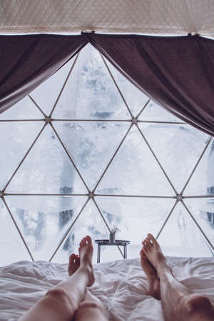 Legs of a man and woman couple in bed against the background of a snow-covered forest in a dome...