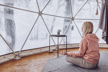 A woman sits in a geo dome glamping tent and meditates, does pranayamas, looks at the winter snow-covered nature.の写真素材