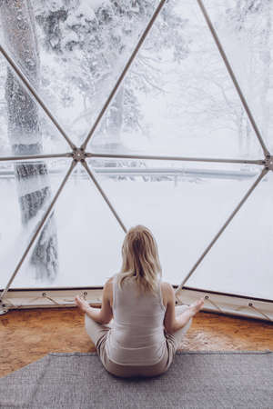 A woman sits in a geo dome glamping tent and meditates, does pranayamas, looks at the winter snow-covered nature. rear viewの写真素材
