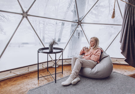 A woman is sitting in a geo dome glamping tent in a bag chair and looking out at the winter snow-covered nature and drinks coffee. Glamping vacation lifestyle concept.の写真素材