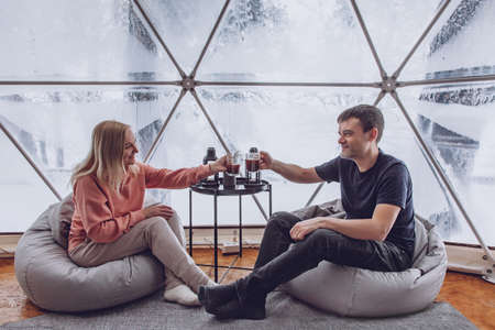 Couple - man and woman are sitting on bag chairs and clinking cups of coffee in geodome glamping...