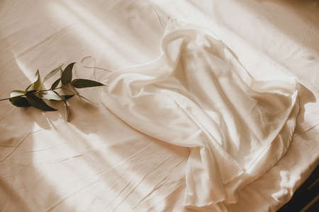 A silk white top with thin straps lies on a crumpled sheet with a branch of eucalyptus in the...