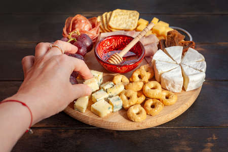 Womans hand takes a piece of cheese from the Appetizers board with assorted cheese, meat, sausage rosette, grape and cookies. Charcuterie and cheese platter. Top viewの写真素材