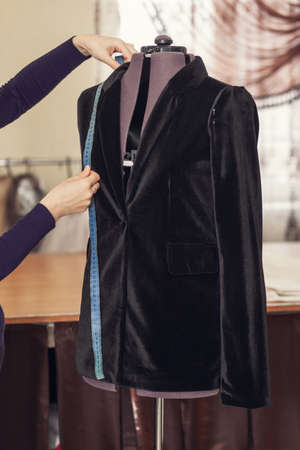 A female dressmaker sews and measures details on a black corduroy jacket hanging on a mannequin in her atelier, close-upの写真素材