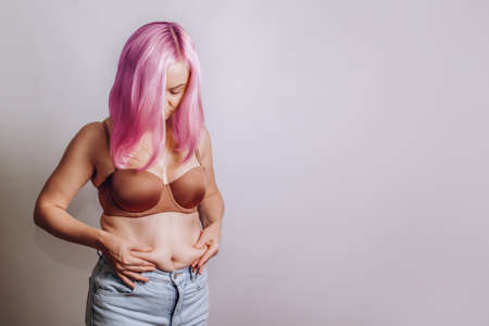 Woman with pink hair squeezes belly fat on her stomach. Woman worries that she is getting fat. Space for text, copy spaceの写真素材