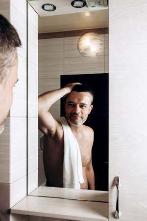 A man looks in the mirror in the morning and straightens his hair in the bathroom with a towel on...