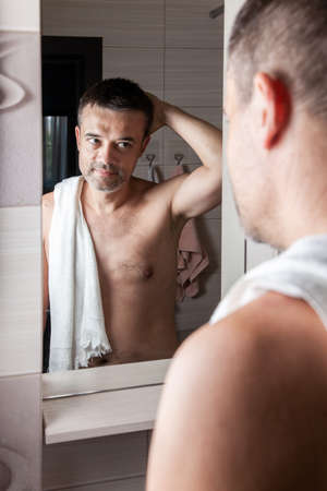A man looks in the mirror in the morning and straightens his hair in the bathroom with a towel on his shoulder.の写真素材