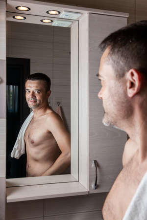 A man looks in the bathroom mirror in the morning with a towel on his shoulder.の写真素材