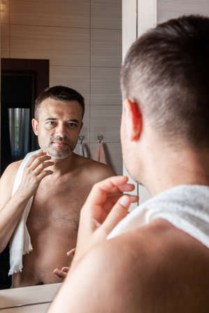 A man looks in the mirror and touches his chin with stubble, going to shaveの写真素材