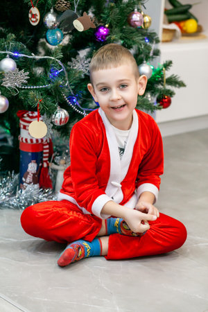 The boy is sitting under the Christmas tree in a Santa costume without a hat, looking at the camera and smiling.の写真素材