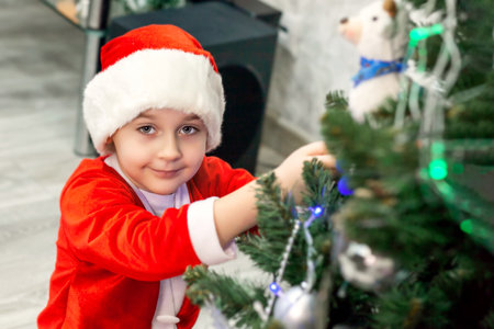 A boy in a Santa costume is decorating a Christmas tree, looking at the camera and smiling.の写真素材