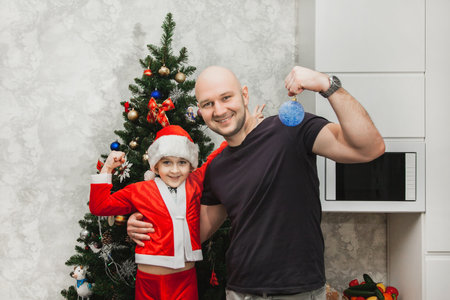 Dad and son in a Santa costume are standing near the Christmas tree and showing biceps, looking into the camera.の写真素材