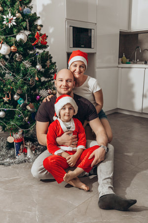 Family - mom, dad and son in Santa costume sitting under the Christmas tree on the floor in their new apartment, looking into the camera.の写真素材