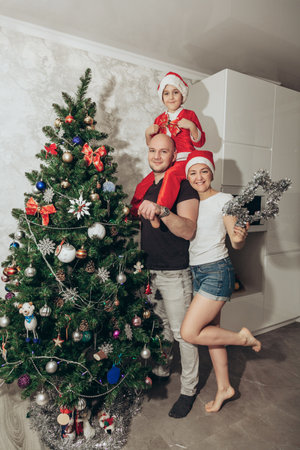 Family mom, dad and son in Santa costume fool around and play near the Christmas tree in their apartment, looking into the camera.の写真素材